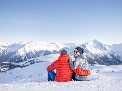 Winter delights in Gerlos • Der Grubacher: Eco Hotel in Gerlos Couple sitting in the snow enjoying the view of snow-covered Alps under a blue sky