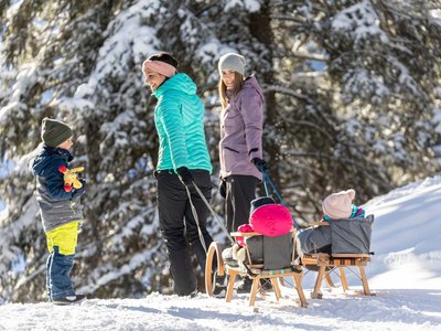 Winter delights in Gerlos • Der Grubacher: Eco Hotel in Gerlos Two women pull children on sleds through a snowy winter forest
